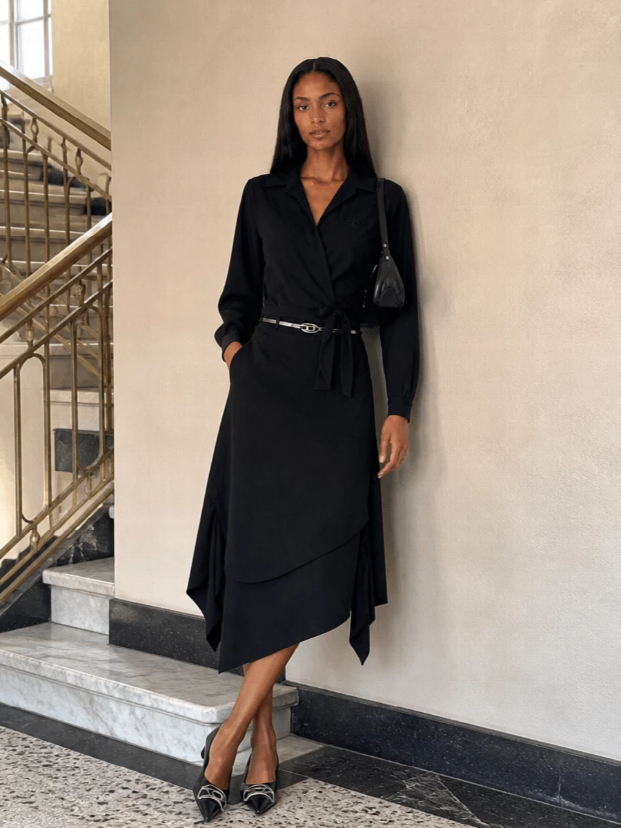 A black-on-black wrap pairing turns the shirt into a sharp, sculpted column, with the asymmetric midi echoing its tied waist for a clean, directional line. Silver-pointed flats, a curved shoulder bag and a slim metallic belt bring just enough hardware shine to read after-hours while staying quietly minimal. The result is a city-ready outfit that moves from office to late cocktails without losing its edge.