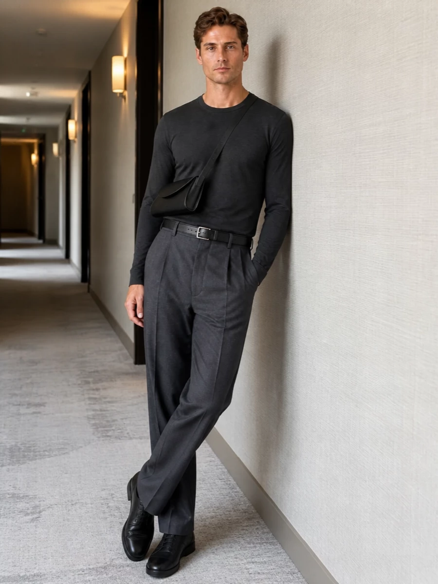 A charcoal long-sleeve tee sits against high-rise pleated trousers in a slightly lighter grey, creating a calm, tonal column. Near-dress black leather sneakers keep the mood precise but relaxed, while a slim black belt and crescent shoulder bag finish the outfit with subtle structure and city readiness. The result is a restrained, quietly confident uniform that moves easily from desk to dinner without ever feeling overworked.
