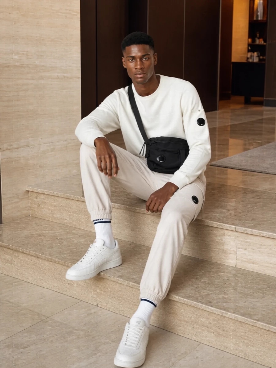 A soft off white C.P. Company crew meets light stone cargos for a clean, technical take on neutral streetwear. Tonal Comme des Garçons sneakers keep the silhouette streamlined while a compact C.P. crossbody and striped socks add just enough utility and graphic detail. The result is a calm, city ready uniform that feels airy but still engineered.