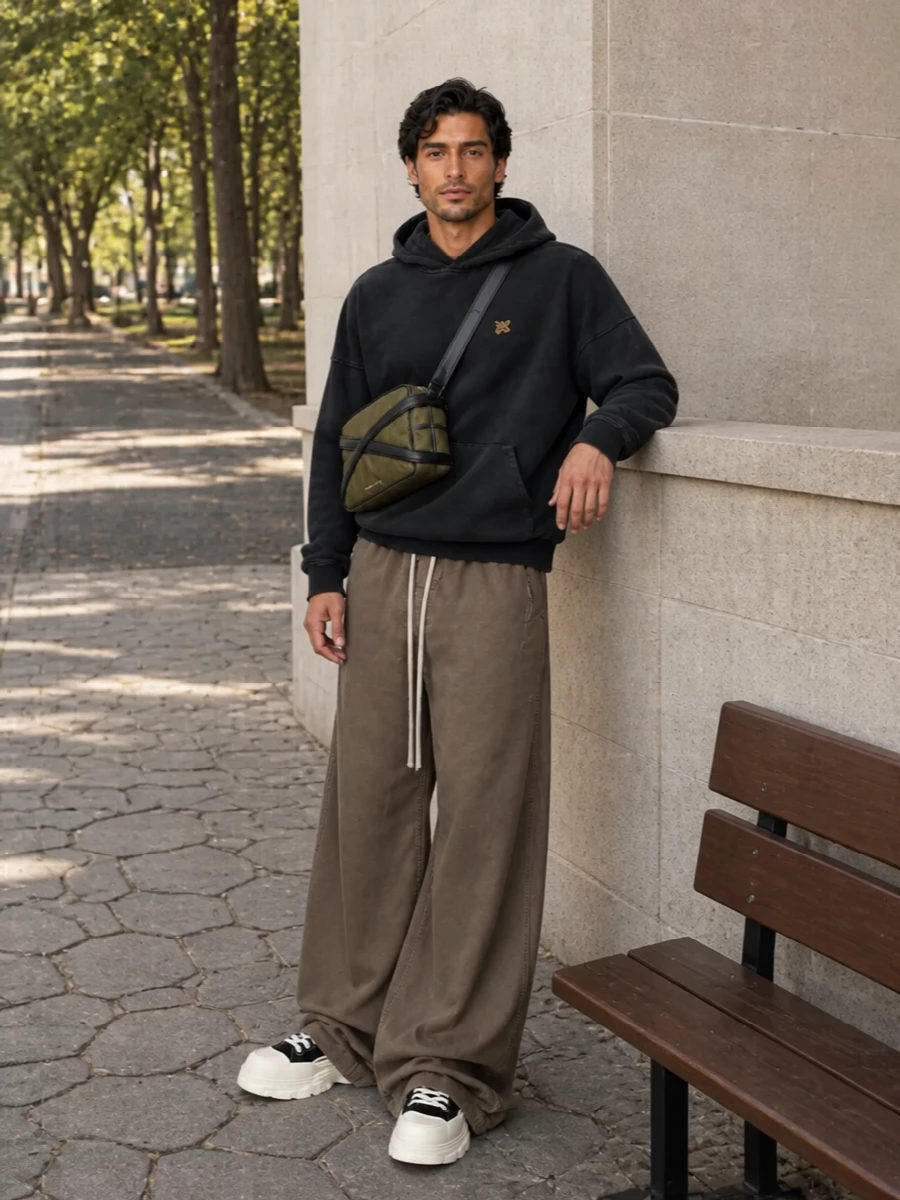 Ultra wide taupe DRKSHDW trousers pool over sculpted black and off white McQueen sneakers for a low-slung, graphic base. A washed black Palm Angels hoodie keeps the top half quiet and slouchy, letting the volume read as intentional. The structured khaki crossbody adds a small hit of utility color and function without disturbing the dark, minimalist street palette.