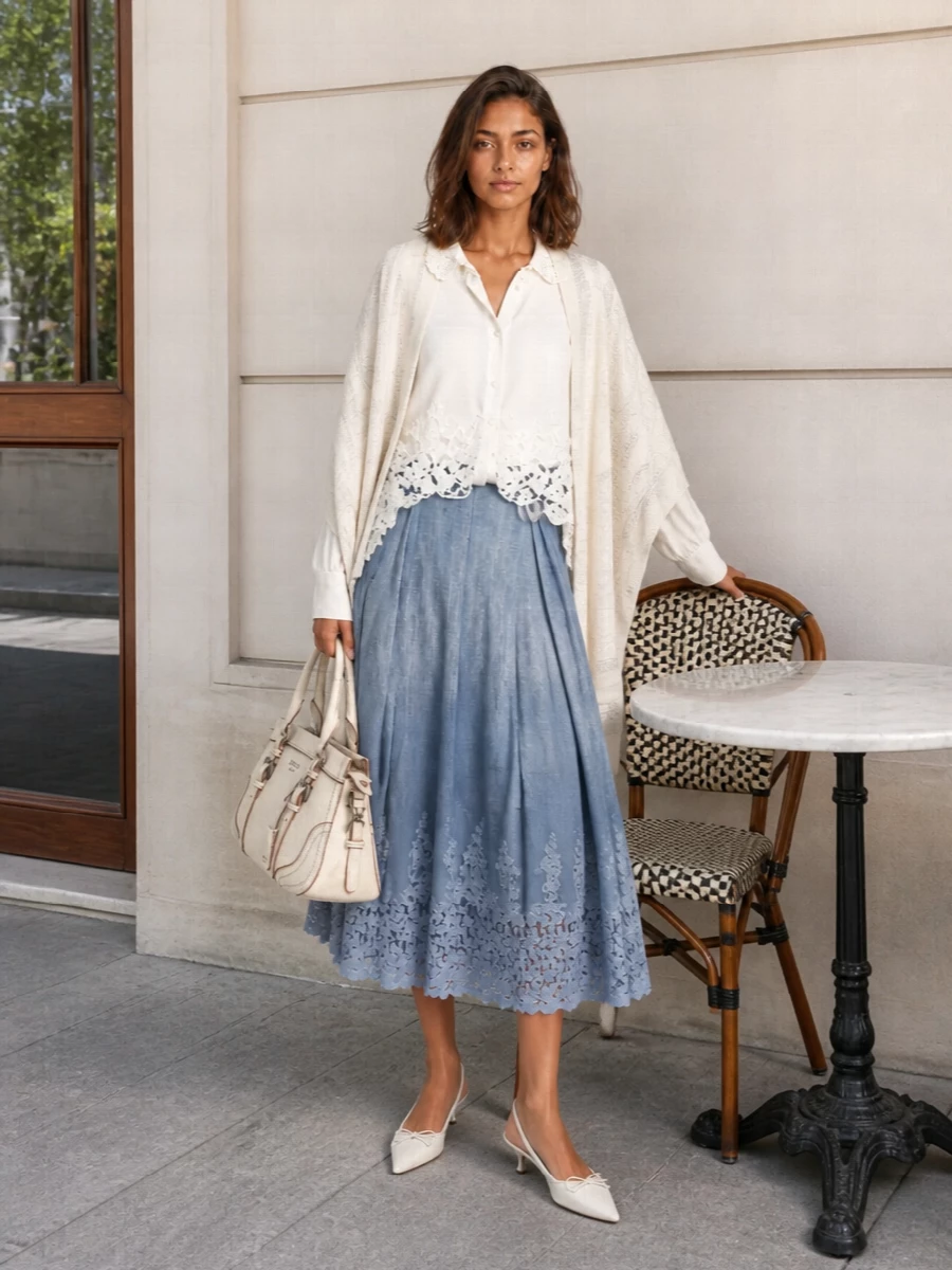A soft ivory lace-trim blouse tucked into the dusty blue pleated midi skirt creates a romantic, elongated line that feels composed yet fluid. White kitten-heel slingbacks and a structured cream top-handle keep the palette light and formal enough for lunches that drift into elegant evening plans. A tonal paisley stole adds a subtle layer of texture and warmth without disturbing the serene harmony of blues and creams.