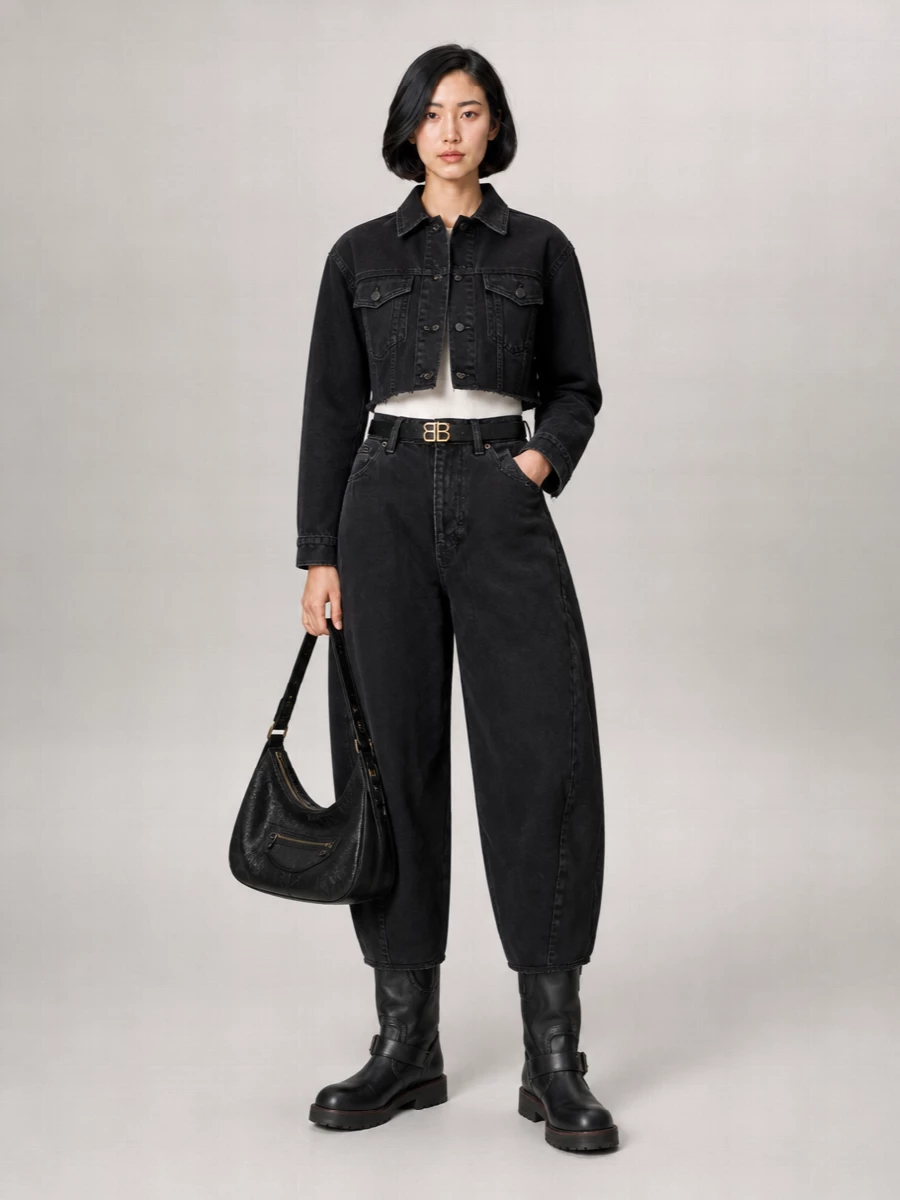 Cropped black denim up top and sculptural curved-leg jeans create a strong, high-waisted line that feels distinctly Balenciaga, all in a deep washed-black palette. Chunky moto boots and a slouchy black shoulder bag add weight and texture, giving the uniform a tough, street-grounded finish. A slim logo belt sharpens the waist so the volume reads intentional rather than oversized.