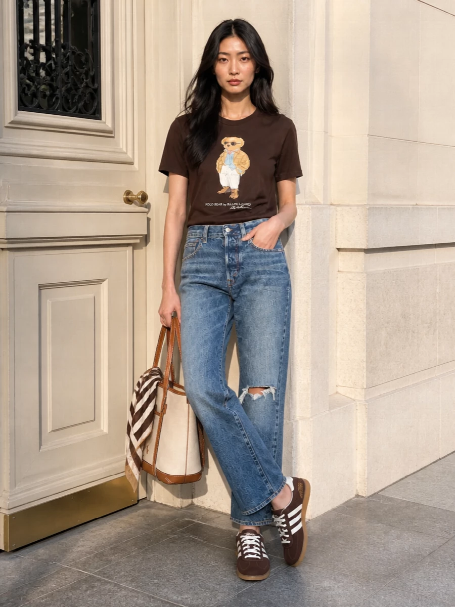 Chocolate Polo Bear, mid blue denim and brown suede sneakers create a laid back, heritage-sport mix that still reads considered. The straight leg keeps the proportions easy around the graphic tee, while the canvas-and-leather tote and striped scarf sharpen the palette in warm neutrals. It is an easy city uniform for coffee runs, casual Fridays and weekend wandering.