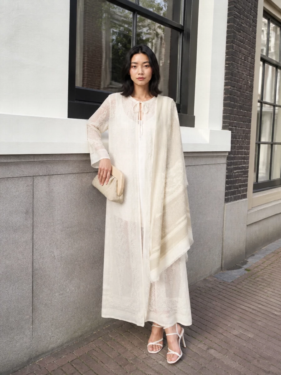 A long, sheer ivory dress meets ultra-fine ivory strappy heels for a column of light that feels both modern and quietly romantic. The buttercream framed clutch and tonal paisley stole bring just enough texture and interest, keeping everything in the same soft neutral register. It is an understated summer evening ensemble that reads serene, elongated and intentionally luxurious.