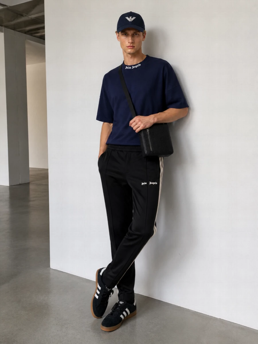 A navy Palm Angels tee and black striped track pants create a clean, athletic base that feels modern and relaxed under the Emporio Armani cap. Black and white terrace sneakers keep the lines streamlined while the Gucci crossbody brings a quiet hit of luxury to the street-sport mix. The palette stays tight in navy, black and white, so the graphic branding reads sharp rather than loud.