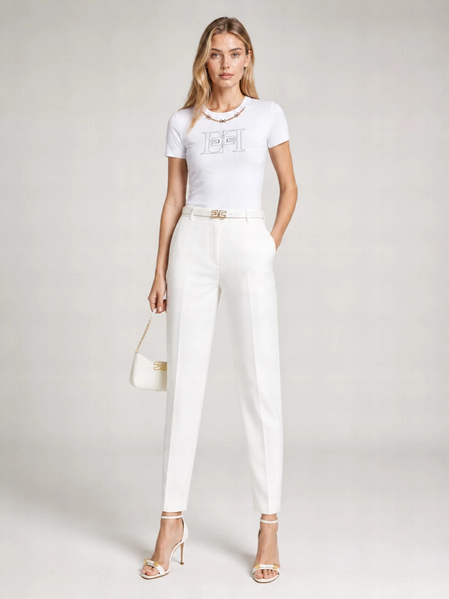 A crisp white-on-white palette lets the chain-detailed tee and high-rise tailored trousers do the quiet talking, all trimmed with discreet flashes of gold. Stiletto sandals and a compact baguette keep the line long and clean, taking this from polished day to luminous summer evening in one move. The slim white belt is the final, almost invisible touch that sharpens the waist without interrupting the minimal mood.