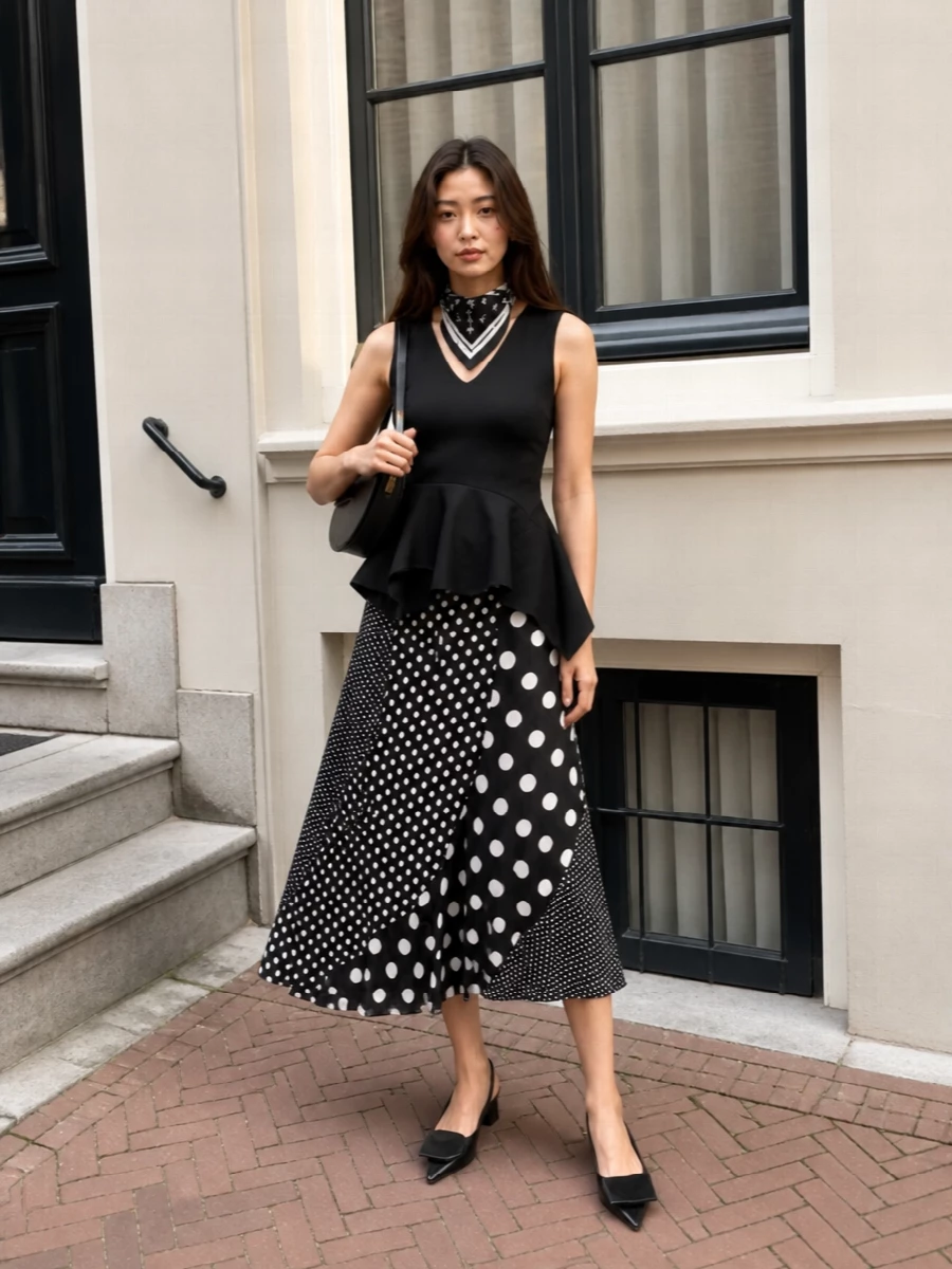 Monochrome polka dots meet sculpted black tailoring for a graphic, romantic outfit that moves with you. The asymmetric peplum echoes the skirt’s sweep, while the pointed slingbacks and curved shoulder bag keep the lines clean and modern. It is gallery-ready but still easy for long city days that drift into dinner.