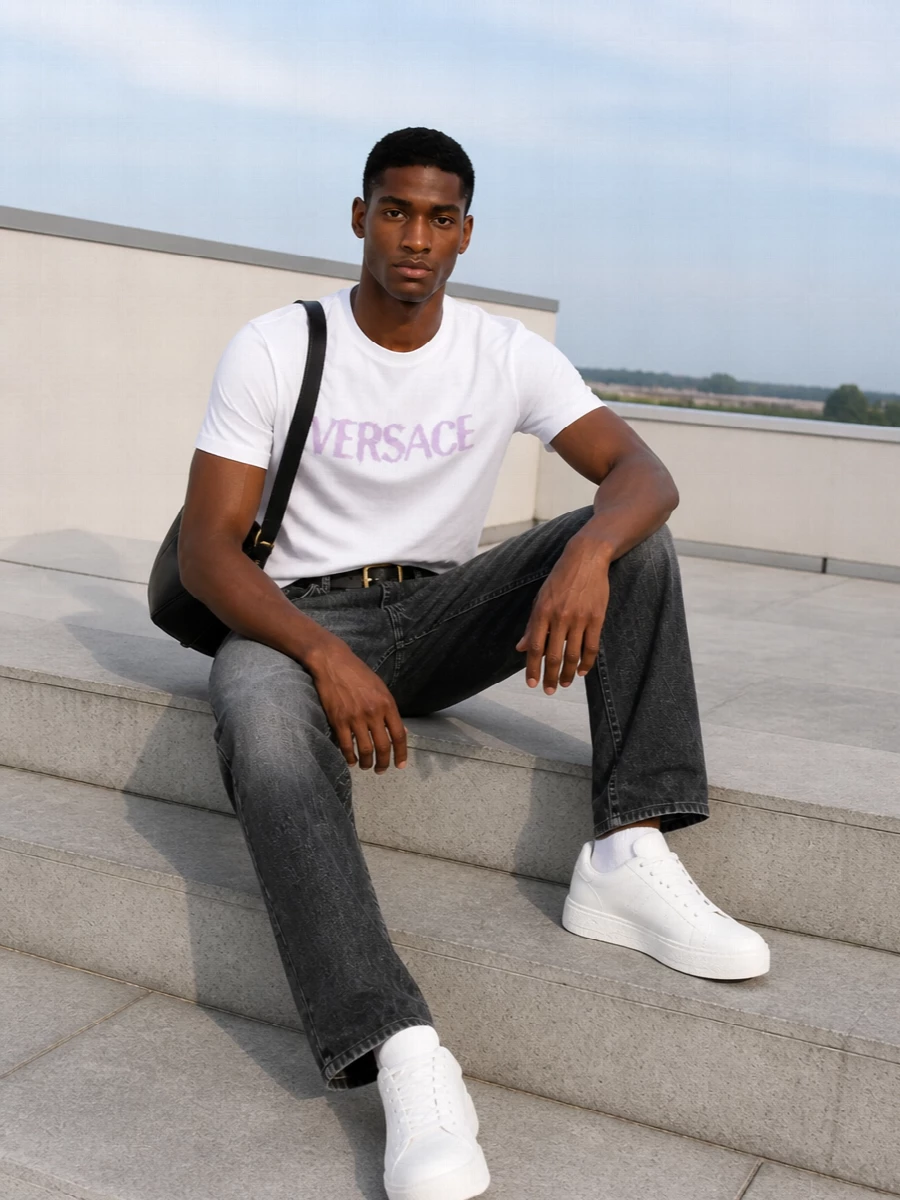 A crisp white Versace logo tee in washed lilac sits against charcoal straight-leg denim for a clean, modern take on branded streetwear. Tonal white leather sneakers keep the line light, while a black leather shoulder bag and slim belt introduce just enough hardware for a polished city finish. It is an easy off-duty uniform that feels precise rather than loud, ideal for warm-season days that move from errands to dinner.