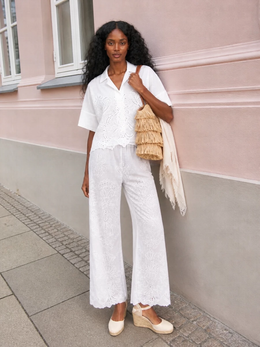 A head-to-toe white story keeps the eye on texture, with broderie and sheer embroidery creating a soft play of light over the relaxed wide-leg silhouette. Espadrille wedges lengthen the line while a fringed raffia bag and blush-toned scarf bring just enough bohemian warmth for garden parties and coastal lunches. The result feels romantic, airy and quietly polished for warm summer days.