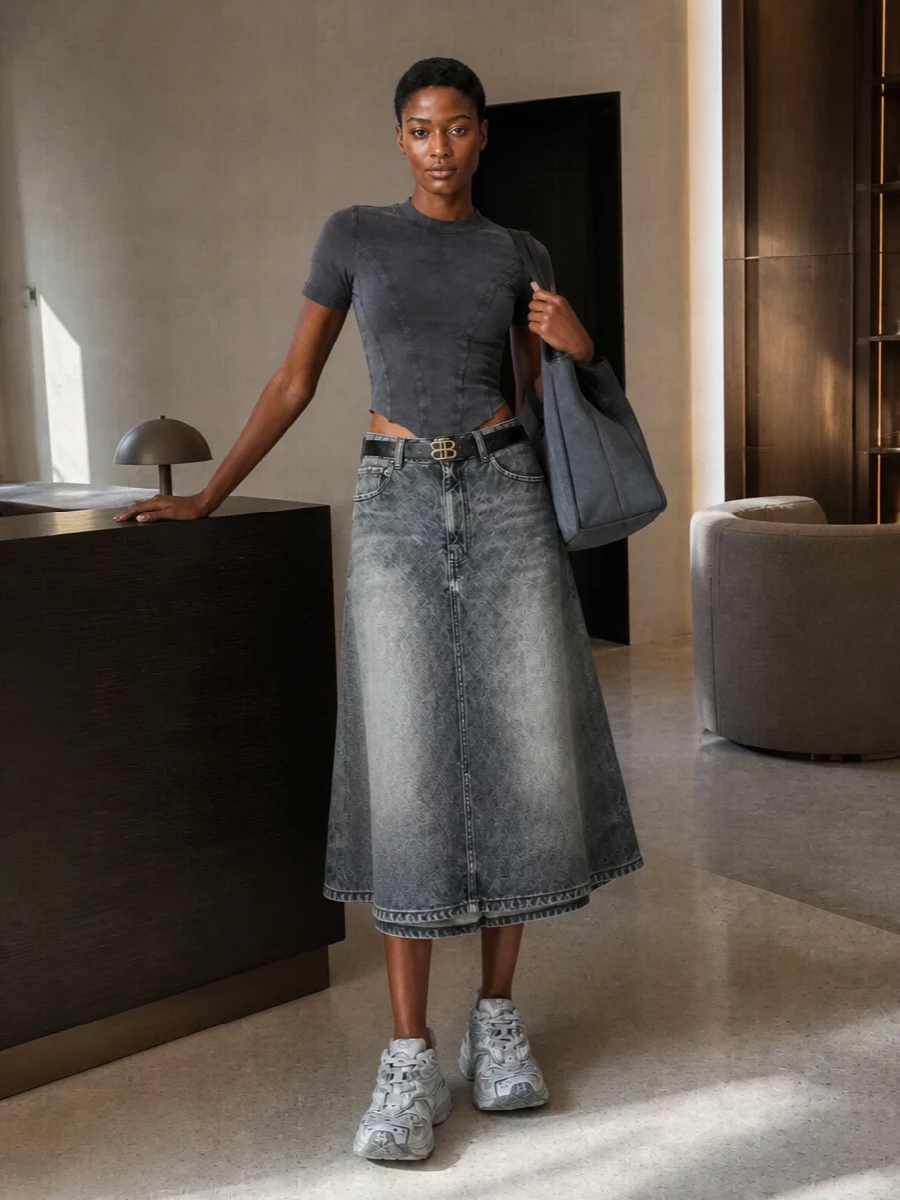 A washed-charcoal corset tee sharpens the A-line denim midi, giving you a clean waist over that easy flare. Chunky grey sneakers keep it grounded and relaxed, while a large charcoal tote and slim logo belt add just enough structure for city days that stretch into late nights. It is pure Balenciaga street, all in tonal greys that feel quietly directional rather than loud.