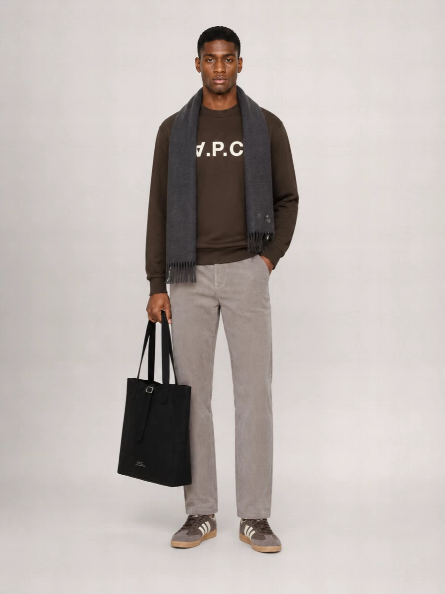 A.P.C.’s dark brown logo sweatshirt sits over taupe cord trousers for a calm mix of depth and texture. Brown and cream terrace trainers echo the logo hit, keeping everything grounded yet relaxed. A structured black tote and charcoal scarf finish the city-ready uniform with clean, understated utility.