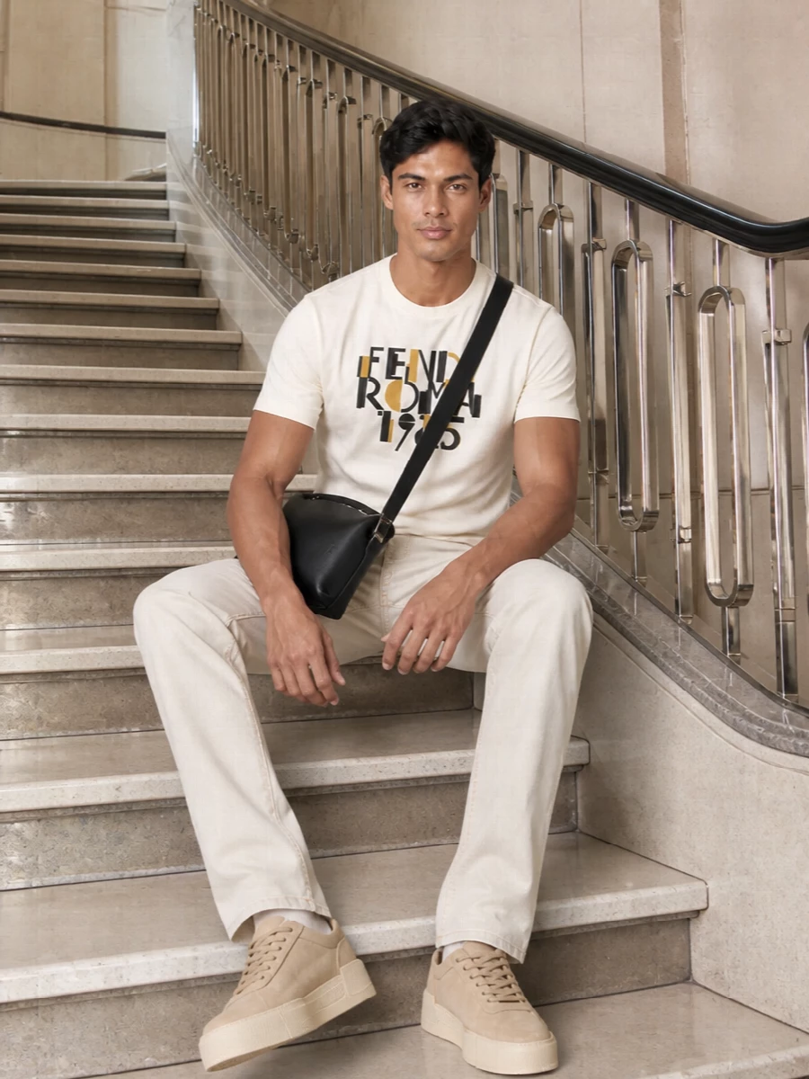 A buttercream Fendi graphic tee sits over stone Versace denim for a light, neutral base that still reads considered. Sand-suede Fear Of God sneakers keep everything tonal and modern, while a black Loewe crossbody quietly picks up the tee’s graphic black accents. The result is relaxed, city-ready and quietly luxurious without feeling over-styled.