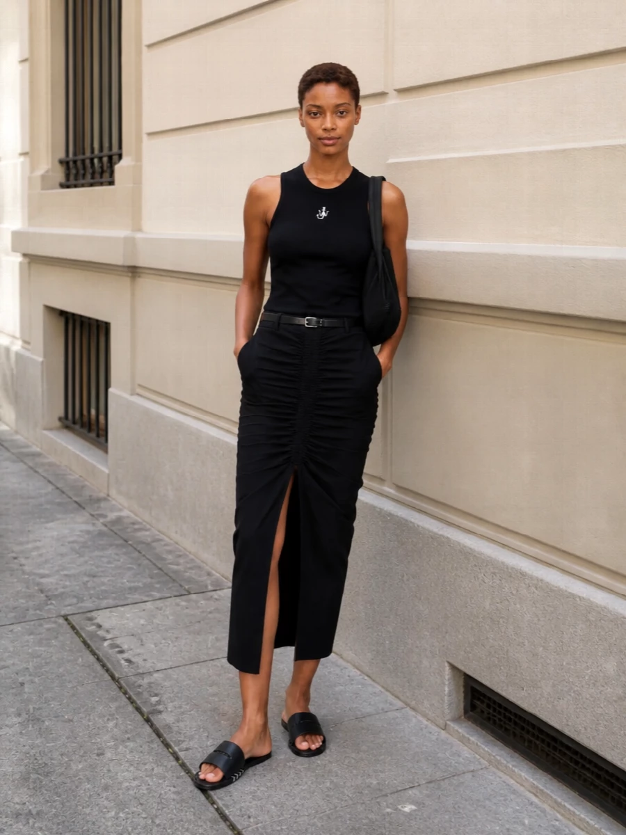 A sculpted black tank meets a ruched midi skirt for a long, pared-back line that feels quietly assertive. Black slides and a soft shoulder bag keep the mood urban and unfussy, while a slim belt adds just enough structure at the waist. It is an easy day-to-night uniform that reads considered without trying too hard.