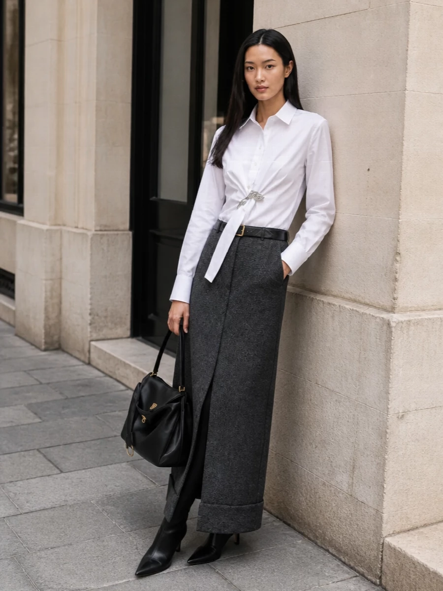 This look centers on the charcoal herringbone maxi skirt and the asymmetric white Prada shirt, creating a refined FW column of tailoring with a subtle, sculptural twist at the waist. Pointed black Prada ankle boots and a structured Balenciaga top‑handle bag sharpen the silhouette further, while a slim black quilted belt quietly defines the waist for a composed, modern evening‑ready ensemble.