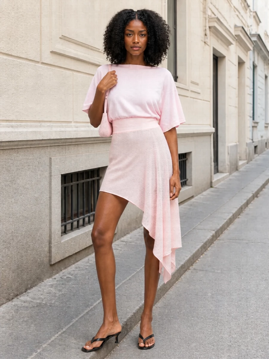 A blush-on-blush knit set lets the asymmetric mini feel soft and sculpted at once, with the matching top framing the waist and the drape. Sleek black kitten-heel mules cut through the sweetness just enough, while a woven pink baguette and whisper-light wrap keep the mood romantic and ready for long summer afternoons that stretch into evening. The result is relaxed, feminine polish with a quietly graphic edge at the foot.