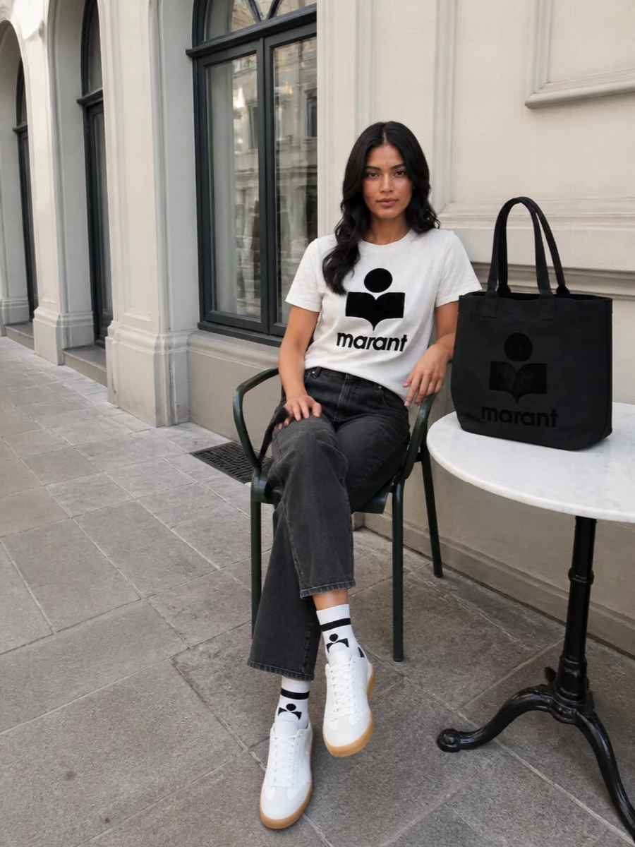 Graphic logo tee, washed charcoal denim and crisp white sneakers create an easy Parisian streetwear uniform with a clear black and off-white story. The straight-leg jeans keep the line relaxed but intentional, while the oversized black tote and matching logo socks pull the branding into a cohesive, city-ready finish.
