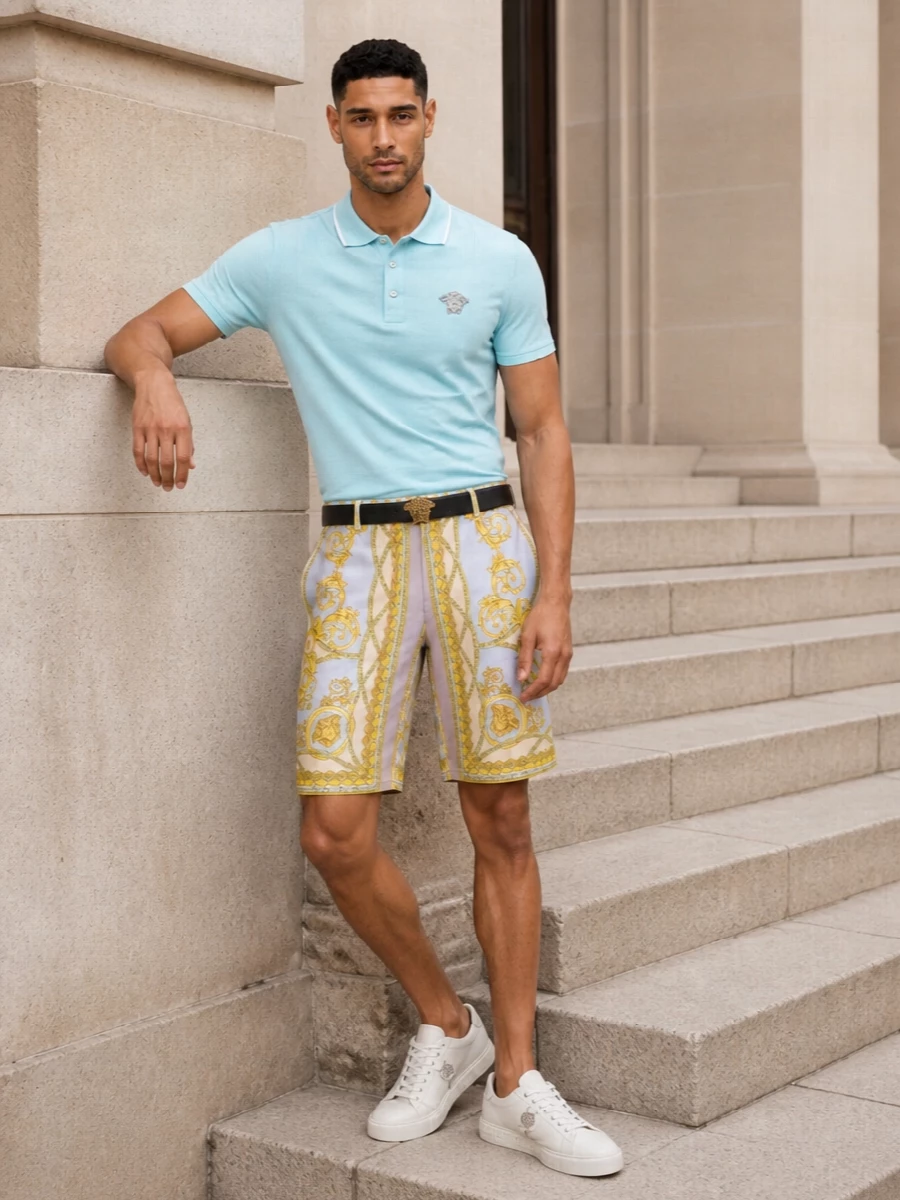 Versace’s baroque shorts take the spotlight, framed by a soft aqua polo that echoes the blue in the print without competing with it. Clean off‑white sneakers and a structured pale blue tote keep the mood modern and graphic, more resort-city than costume. A sharp black Medusa belt gives just enough gold hardware at the waist to connect the whole palette and underline the house attitude.