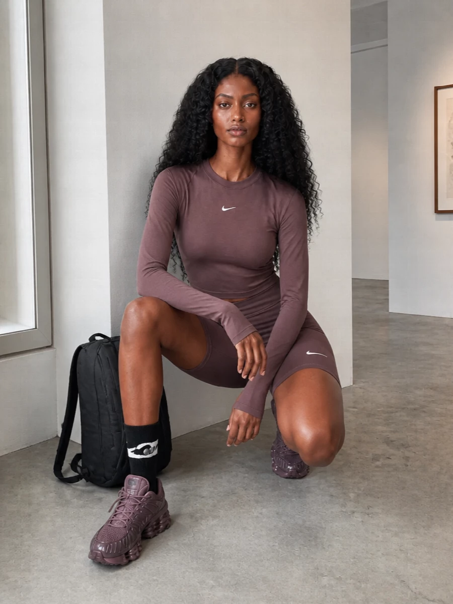 A monochrome plum Nike set anchors this outfit with a clean, body-hugging line that feels ready for the studio or a long city walk. Sculpted runners in a matching tone and a dark technical backpack keep the vibe streamlined and performance minded, while graphic crew socks add just enough detail at the ankle. The result is tight, modern and quietly bold, all built around that rich brown-plum base.