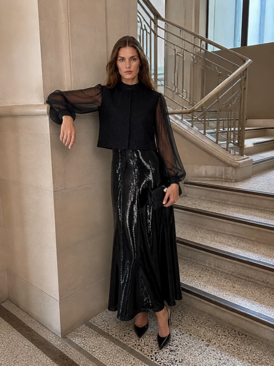 I’m styling the cropped Armani jacket over the Alberta Ferretti sequin slip gown, creating a long, liquid black column topped with sheer, jewelry-like sleeves. The Jimmy Choo crystal-slingback pumps and satin bow clutch keep all the sparkle concentrated in small, precise accents so the silhouette stays clean and the mood is modern cocktail. I chose the dress path rather than tailoring because the jacket already behaves like evening suiting, and pairing it with a gown avoids a clash of lapels and multiple tailoring layers.