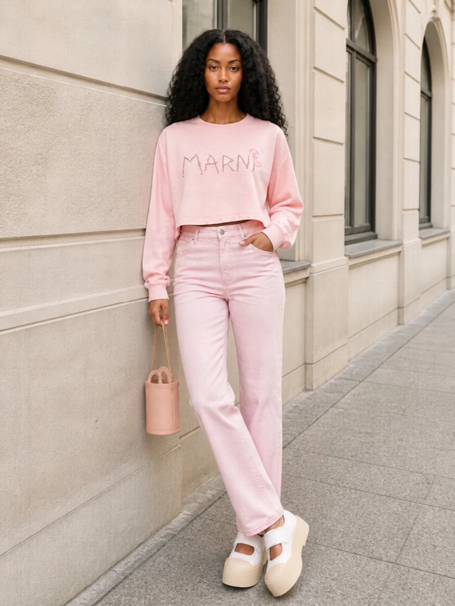 Soft blush tones run from the cropped logo sweatshirt into clean, straight-leg trousers for a column of color that still feels easy. Chunky white Mary Jane sneakers and a tiny blush crossbody keep it playful and polished, leaning into sporty prep rather than full athleisure. The result is a relaxed city-day outfit that feels intentional without losing its charm.