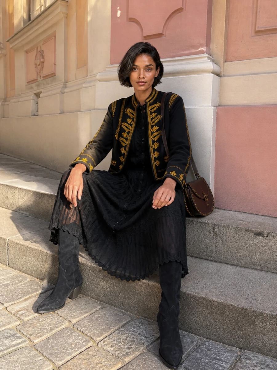 I am styling the embroidered navy jacket with a single hero dress rather than denim, so the ornate gold trim reads deliberate and polished against an inky, romantic base. The black embroidered midi and knee-high suede western boots extend the jacket’s band-inspired, boho energy into a sleek column, while the dark chocolate saddle bag quietly ties together all the warm hardware tones. The result is an evening-ready outfit that still feels grounded and easy to wear from gallery nights to late dinners.