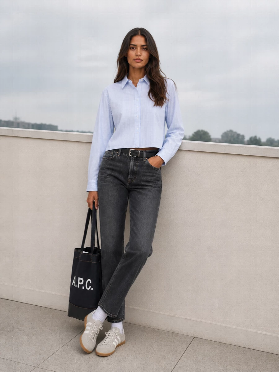 A straight, dark-wash jean anchors an easy mix of washed blues and charcoal, kept fresh with a cropped striped shirt that lightens the palette. Low-profile grey trainers and a structured navy tote keep the vibe clean and urban, while a slim black belt subtly defines the waist so the proportions stay sharp, not slouchy. The result is relaxed city denim that still feels considered enough for all-day wear.