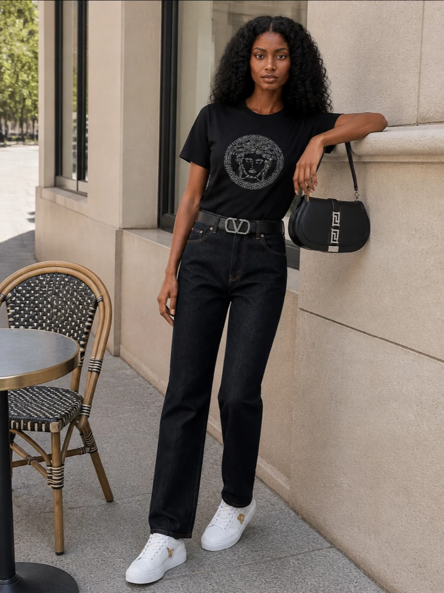 A crystal Medusa tee meets clean dark Versace denim for a polished take on streetwear that feels easy but considered. White Medusa-detailed sneakers keep the line relaxed while the curved black shoulder bag and crystal-buckled belt echo the tee’s shine in a quieter way. The palette stays tight in black, indigo and metal, so the glint reads deliberate rather than loud.