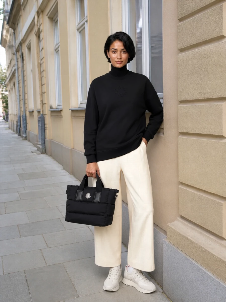A longline black sweatshirt falls cleanly over buttercream wide-leg trousers, creating a graphic yet soft column in quietly athletic fabrics. Cream runners and subtle tipped socks keep the base light and agile, while a padded black tote anchors everything with just enough structure for city movement. The result is a calm, monochrome-neutral pairing that works from airport lounge to low-key dinner without needing a change.