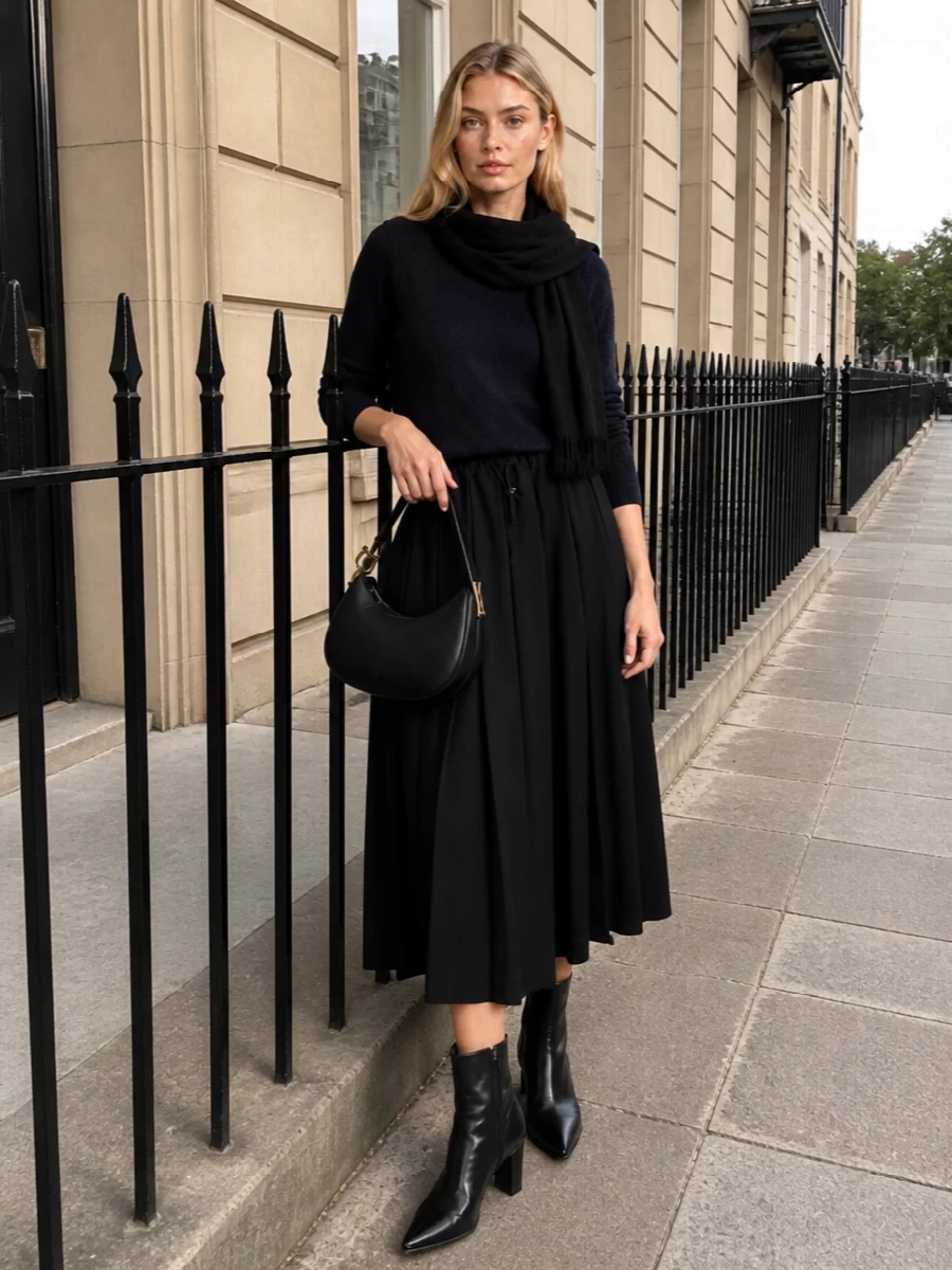 Fabiana Filippi’s black pleated midi skirt is grounded with a midnight navy knit, creating a subtle tonal shift that still reads as pared-back and refined. Prada’s sharp black ankle boots and a compact Ferragamo crescent bag extend the clean vertical line, giving the outfit a quietly assertive, city-ready finish. A gauzy black scarf adds just enough texture for winter without breaking the minimalist rhythm.