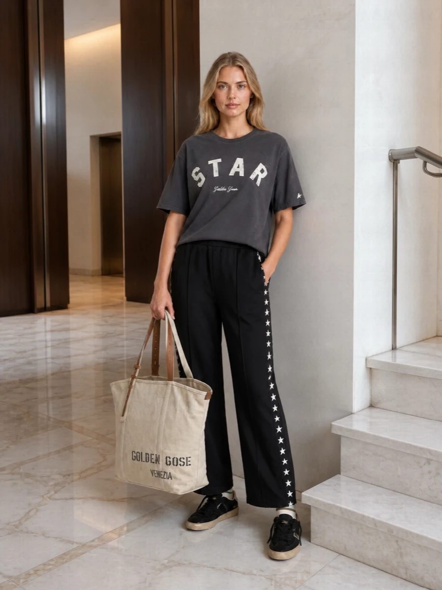 Golden Goose track pants meet a washed charcoal STAR tee for a clean, graphic take on off‑duty dressing. Black suede sneakers keep the line grounded and streamlined, while the oversized canvas tote and striped socks add a relaxed, city‑ready attitude without competing with the minimal palette.