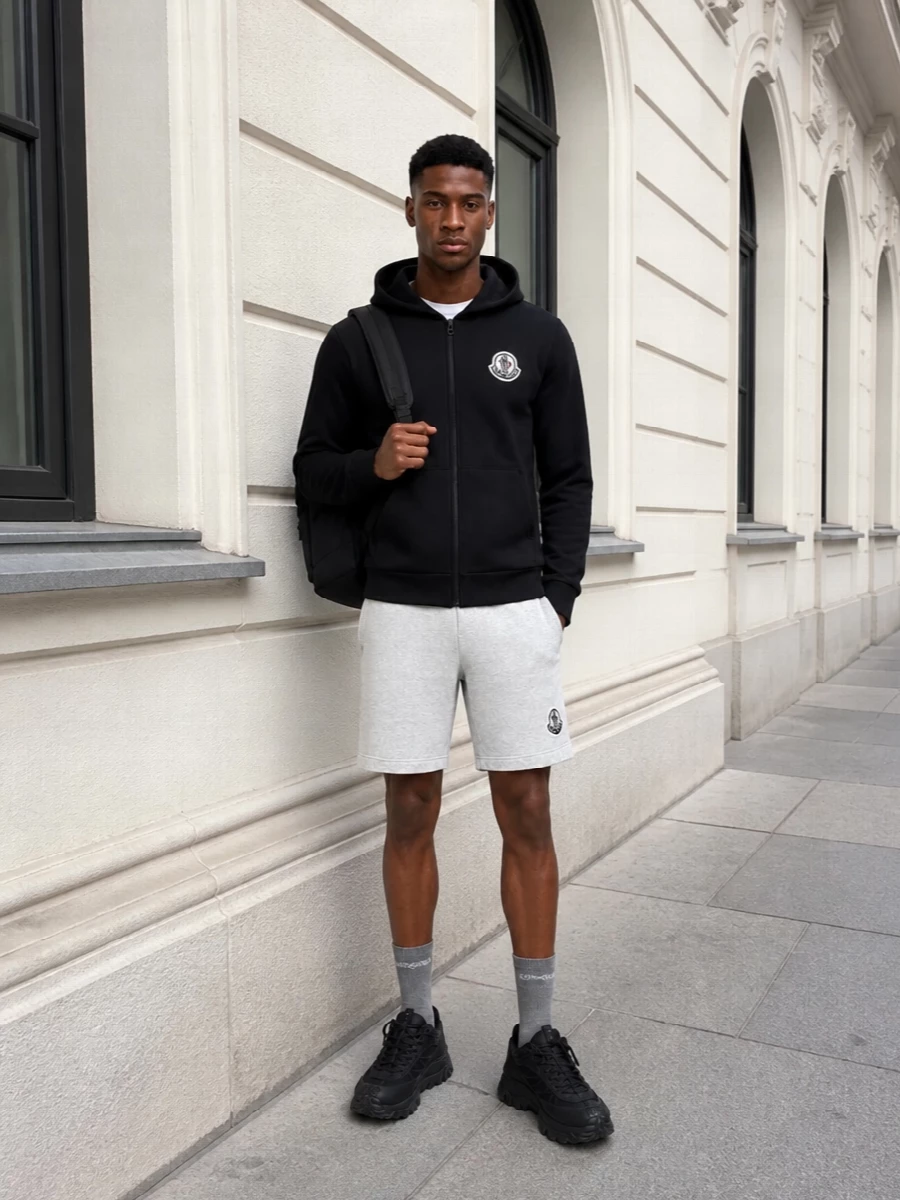 Light heather-grey Moncler shorts are anchored with a black zip hoodie for a sharp, logo-led monochrome contrast. Chunky black sneakers and a matte black daypack keep the energy athletic and urban, while heather-grey socks echo the short’s tone for a clean, considered finish.