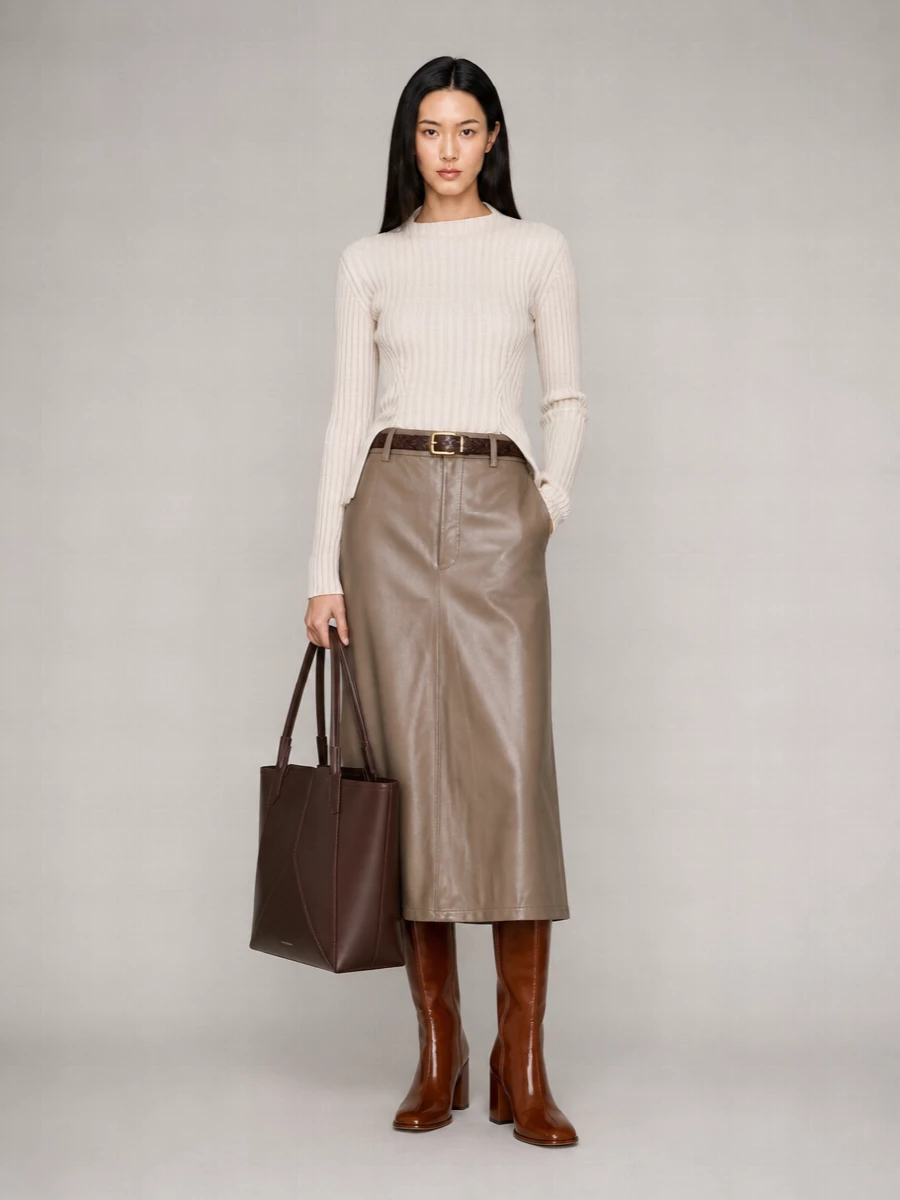 Victoria Beckham’s taupe leather-look midi skirt pairs with the brand’s oatmeal ribbed knit for a soft, tonal take on winter tailoring. Cognac knee-high boots and a chocolate structured tote deepen the neutral palette, keeping the silhouette elongated and quietly polished. A slim dark-brown belt sharpens the waist so the A-line skirt reads clean and intentional from desk to dinner.