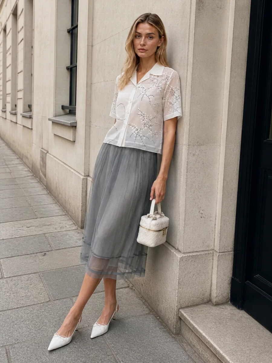 The sheer embroidered Prada shirt is paired with a softly structured grey overlay skirt, creating a diaphanous, tonal column that feels romantic yet precise. Ivory kitten-heel slingbacks and a compact intrecciato top-handle in matching tones sharpen the look just enough, keeping the mood airy, modern and quietly luxurious for SS city evenings or refined daytime events.