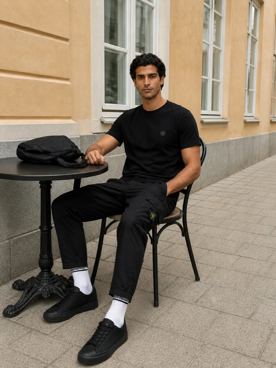 A black Stone Island tee sits over straight black cargos for a clean, technical streetwear uniform that feels ready for the city. All-black sneakers keep the line sharp and unfussy, while off-white sport socks introduce just enough contrast to break up the monochrome without diluting the brand-led mood.