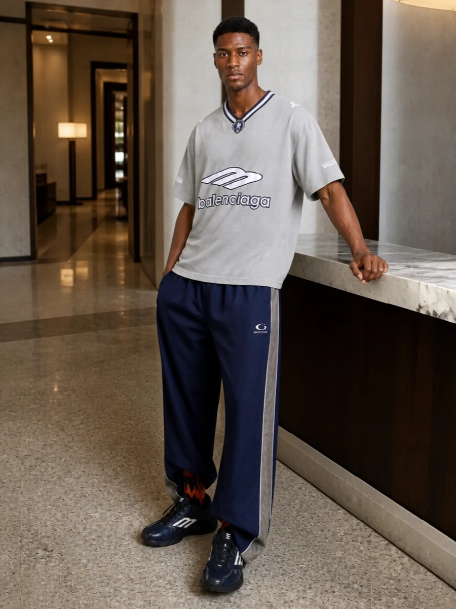 Head-to-toe Balenciaga sport, this pairing leans into the wide navy track pant with a graphic grey jersey top that keeps the palette tight and the energy athletic. Streamlined navy runners keep the silhouette clean and fast, while the bold black duffle and flash of branded socks push it into a confident, arena-to-city uniform. It is relaxed, logo-driven and deliberately coordinated without ever feeling over-styled.