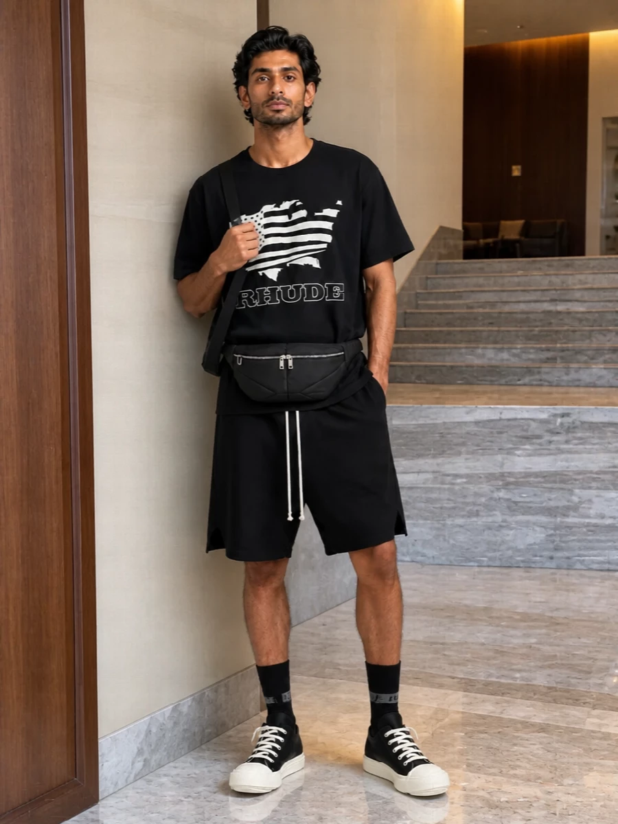 A Rhude graphic tee sits over black Rick drop-crotch shorts for a monochrome, sport-driven street uniform that feels intentional rather than thrown on. Chunky DRKSHDW low-tops echo the tee’s black-and-off-white palette, while a padded belt bag and graphic socks tie the whole mix into a tight, city-ready silhouette. The result is relaxed but focused, with every piece speaking the same high-end streetwear language.