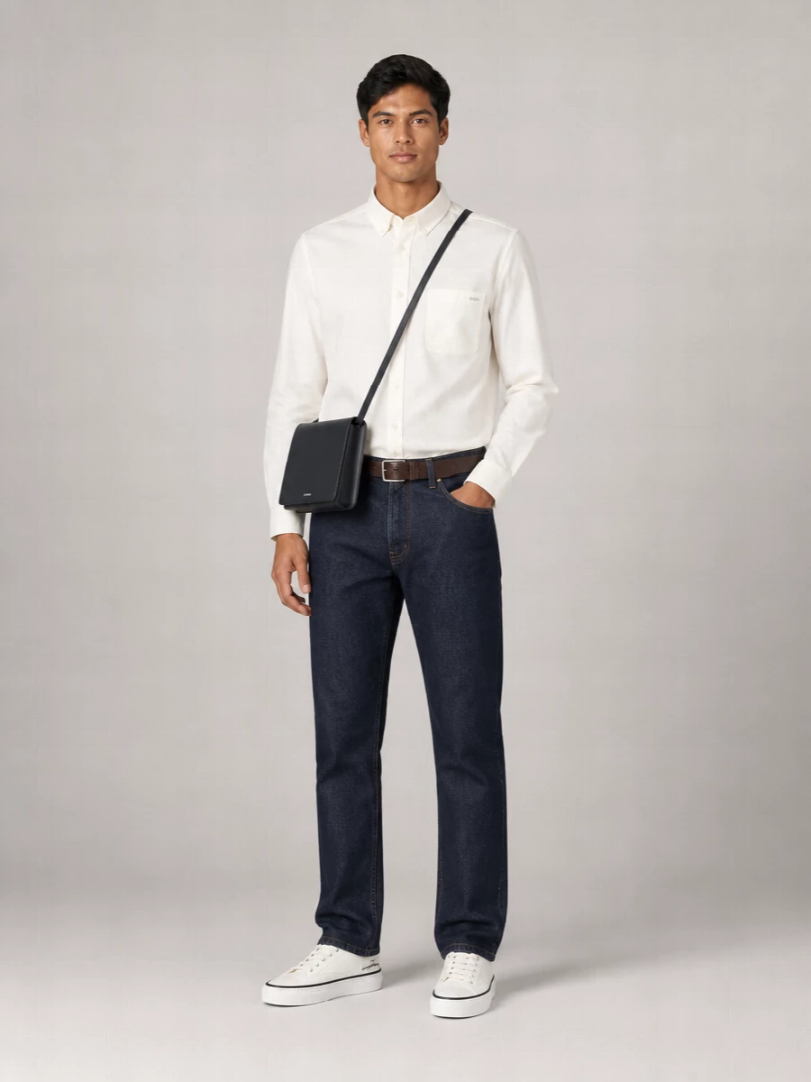Dark indigo denim meets an off‑white button‑down for a clear, considered take on casual dressing that still feels ready for a low‑key dinner. Off‑white canvas sneakers keep the line relaxed but refined, while a deep navy crossbody and brown leather belt add just enough structure and depth without breaking the minimalist palette. It is an easy uniform built on quiet contrasts of blue, cream and leather.
