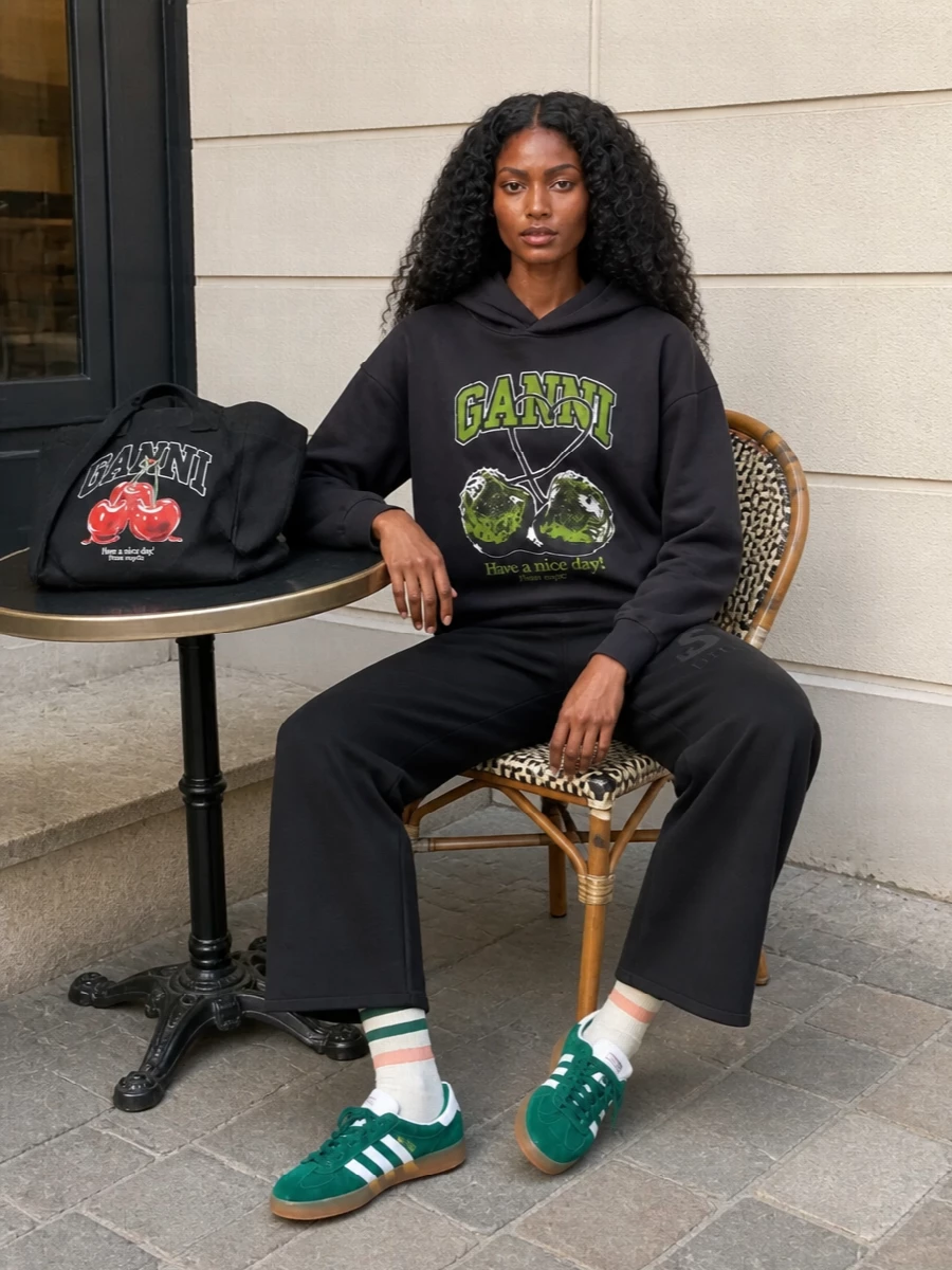 Head-to-toe charcoal and black keep the hoodie’s bold graphic as the focus, with wide-leg sweats reinforcing the relaxed, sport-leaning silhouette. Forest-green Gazelles and the matching Ganni logo tote create a tight color and branding story that feels intentional rather than thrown together. The striped socks are a small but smart link between the footwear and the collegiate mood on top.