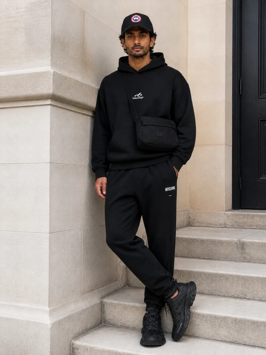 This is a monochrome street-sport uniform built around strong logos and clean performance lines. An oversized black hoodie sits over tapered joggers, then meets aggressively sculpted runners for a stacked, grounded silhouette. The padded shoulder bag adds a low-key technical layer that keeps the all-black palette rich rather than flat.