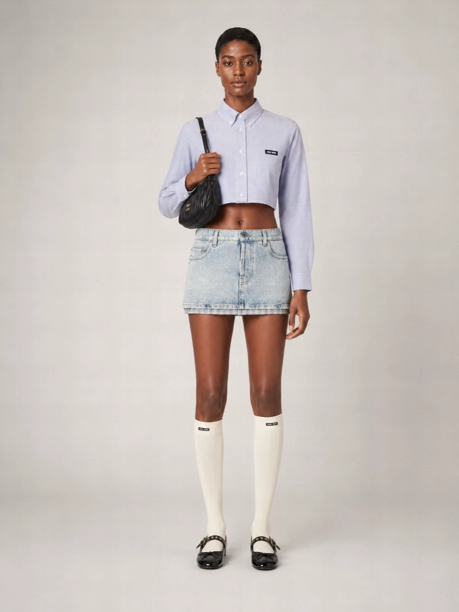 A washed micro denim mini meets a sharp cropped micro-check shirt for a collegiate spin that still feels decidedly rebellious. Black patent Mary Janes and a quilted baguette sharpen the sugariness, while ivory logo socks nod to Miu Miu’s cult schoolgirl styling. The palette stays cool and controlled in blues, greys, and black so the attitude, not the color, does the talking.
