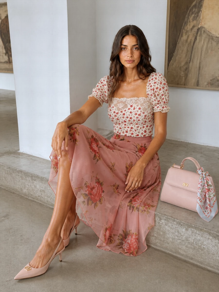 A fitted, lace-trim floral blouse meets a floating dusty-rose chiffon midi for a romantic, Sicilian-garden mood that still feels refined. Blush slingbacks and a structured blush top-handle bag keep the palette soft and polished, letting the florals and delicate neckline stay center stage. A silk floral scarf on the handle or in the hair finishes the story with a light, nostalgic touch.