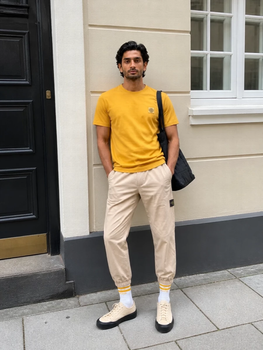 A mustard Stone Island tee sits against light beige technical track pants for a clean, sun-leaned palette that still reads urban and athletic. Neutral mesh-and-suede sneakers and a black technical tote keep everything grounded and functional, while striped socks quietly echo the hit of colour up top. The result is an easy warm-season street-sport uniform that feels considered without losing its relaxed attitude.
