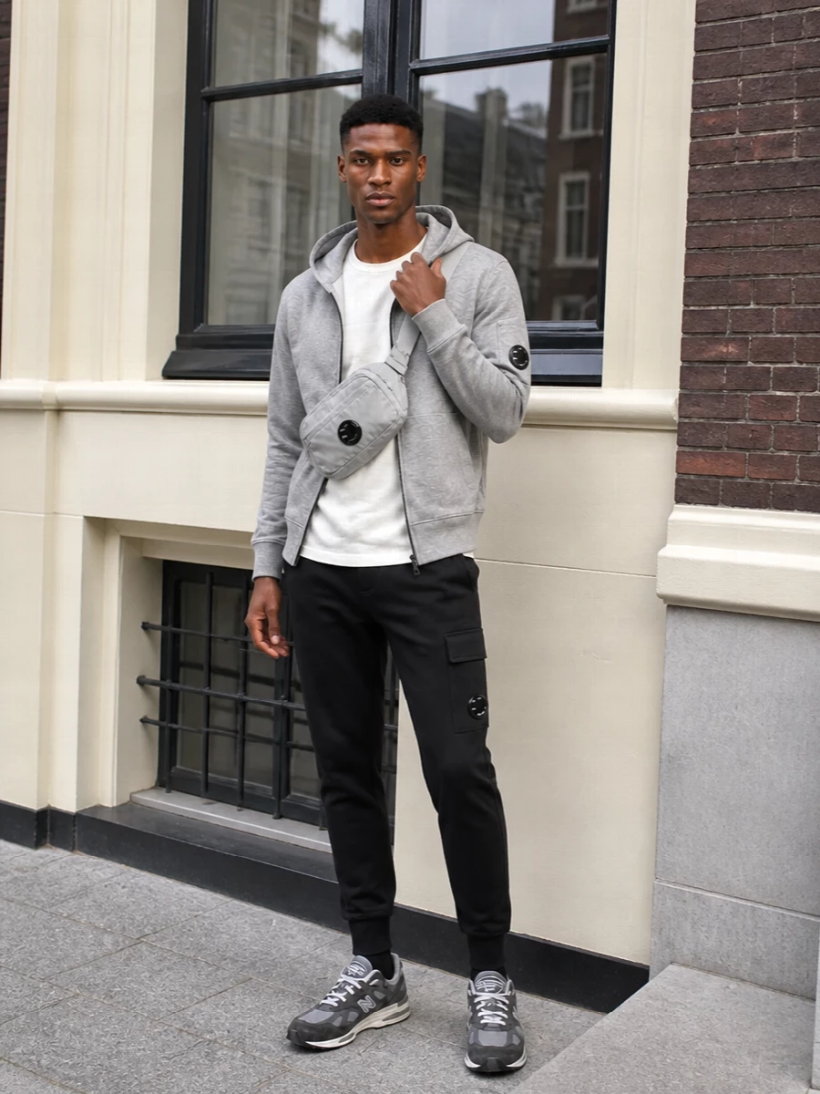 A soft grey and off white C.P. Company story on top sits against sharp black joggers for a clean, technical street mix. New Balance runners keep it cushioned and city ready, while the lens crossbody finishes the utility detail in a tonal way. It is an easy uniform for long days on your feet when you still want the outfit to feel considered.