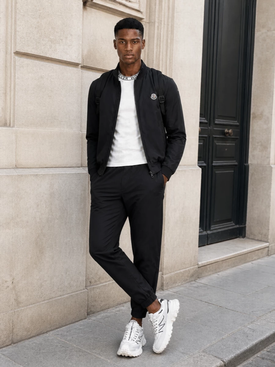 This look is a refined Moncler uniform: a black cropped technical bomber over an off-white logo-collar tee, grounded with tapered black track pants. Off-white technical runners and a pared-back black backpack keep the palette tight and the mood quietly athletic, ideal for spring city days and travel.