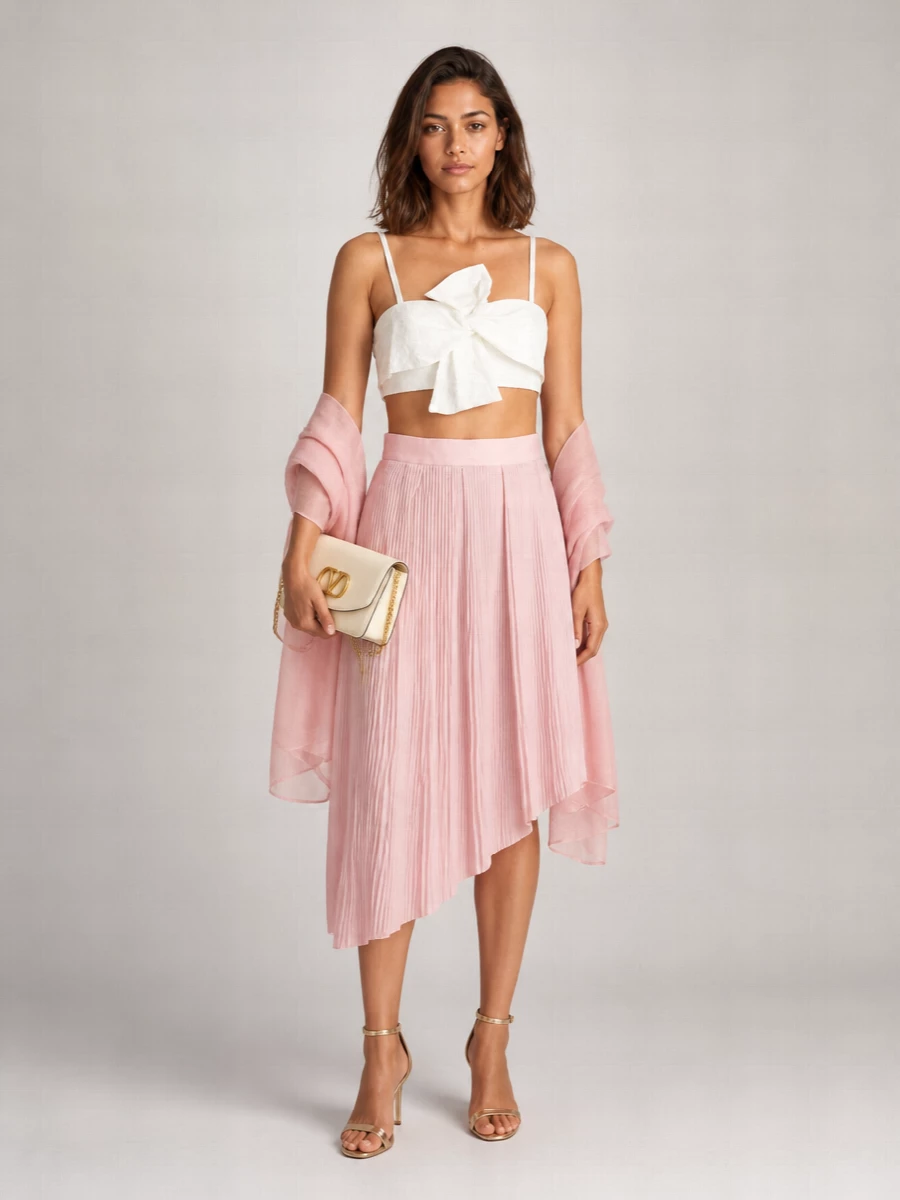 An ivory bow-tied bralette meets a blush pleated midi for a romantic, softly sculpted pairing that feels polished yet light. Champagne-finish stilettos and a buttercream chain clutch bring a quiet shimmer, keeping the palette cohesive and the mood dressy without excess. A gauzy dusty-rose wrap tops it off with a whisper of color and a graceful way to play with coverage after dark.