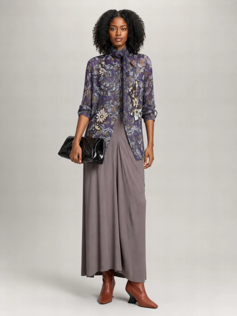 Sheer violet print and jeweled details float over a sculpted taupe maxi skirt, creating a long, romantic line that still reads precise and considered. Rust cone-heel boots and a glossy black clutch keep the Dries mood intellectual rather than sugary, with color held close to the body and hardware kept minimal. The result is an artful evening ensemble that feels dramatic, modern and quietly opulent from gallery opening to late dinner.