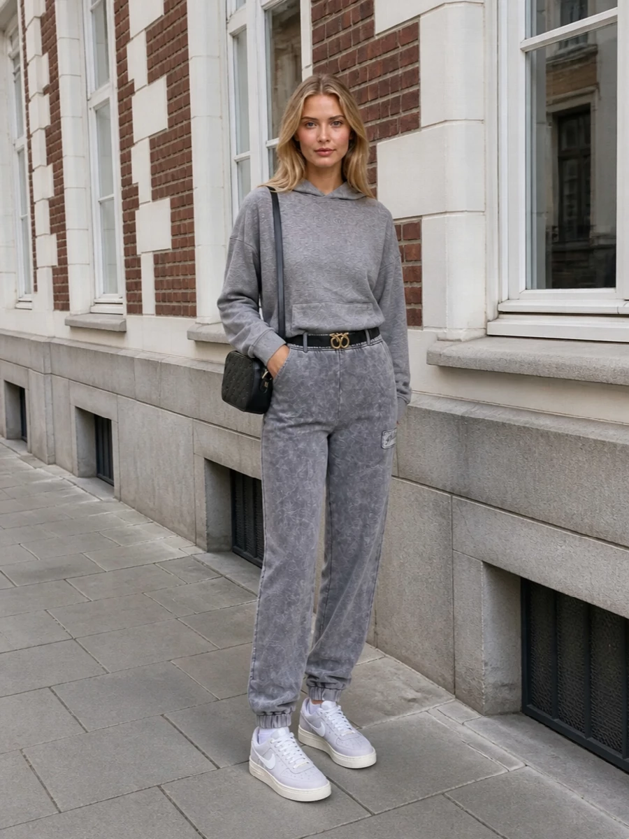 A heather-grey Pinko hoodie pairs with washed-charcoal joggers for a tonal, relaxed set that feels quietly styled rather than thrown together. Soft grey Nike sneakers keep the mood sporty and practical, while a compact black crossbody and optional slim belt introduce just enough structure and polish for city errands or travel days.