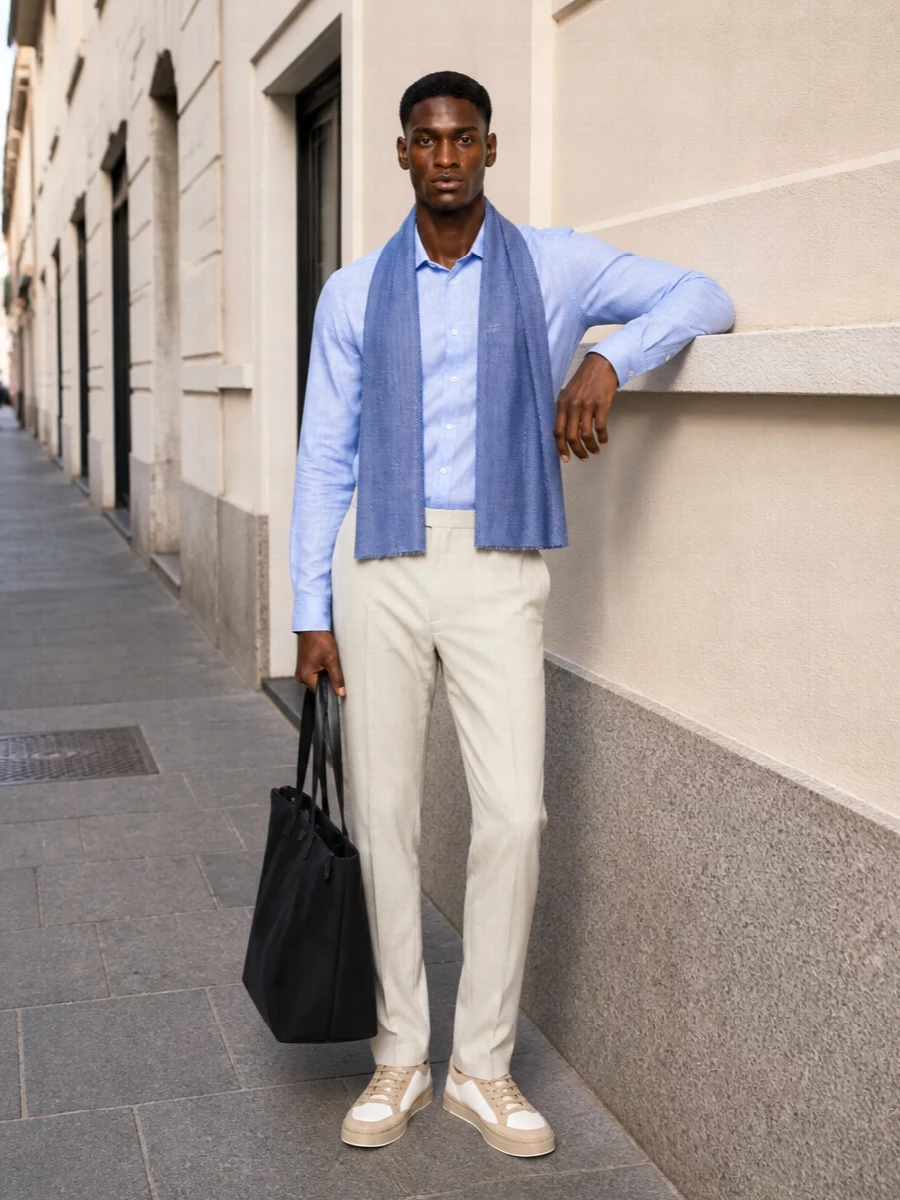 A pale blue shirt with light beige trousers creates a crisp, modern take on smart casual that feels easy in bright weather. Off white and sand sneakers keep the palette soft while the navy tote grounds everything with a discreet, practical note. The blue scarf adds a subtle layer of depth that ties the top half together without breaking the clean lines.
