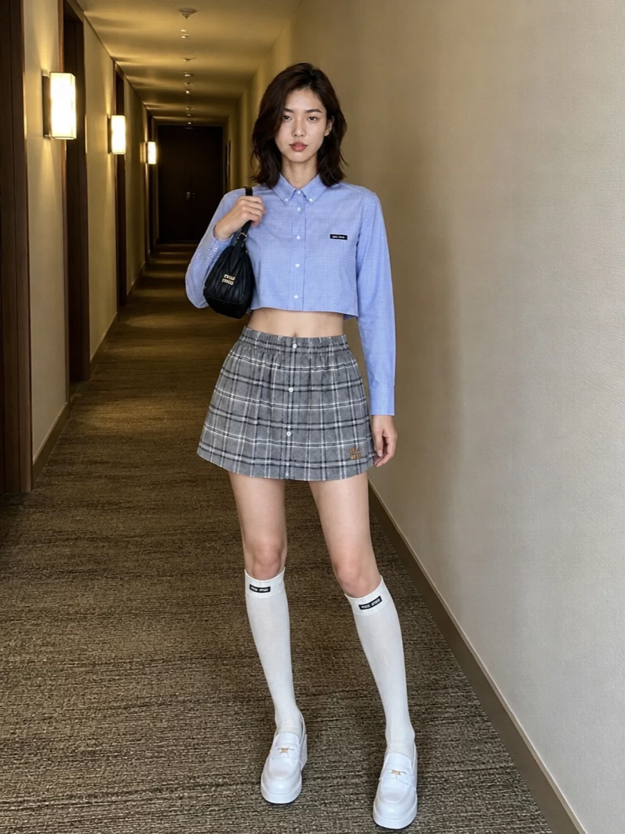 A grey checked mini meets a cropped blue micro-check shirt for a sharp, collegiate take on Miu Miu’s Y2K prep. White platform loafers and logo knee socks keep the leg line long and playful. A quilted black baguette finishes the outfit with compact, polished contrast against the soft neutrals and pale blue checks.