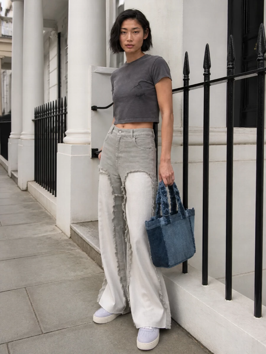 Deconstructed stone-grey denim meets a cropped charcoal graphic tee for a cool, off-duty streetwear moment that still feels intentional. Technical grey sneakers keep the palette tight and practical, while the patchwork denim tote adds capacity and texture without competing with the trousers’ architecture.